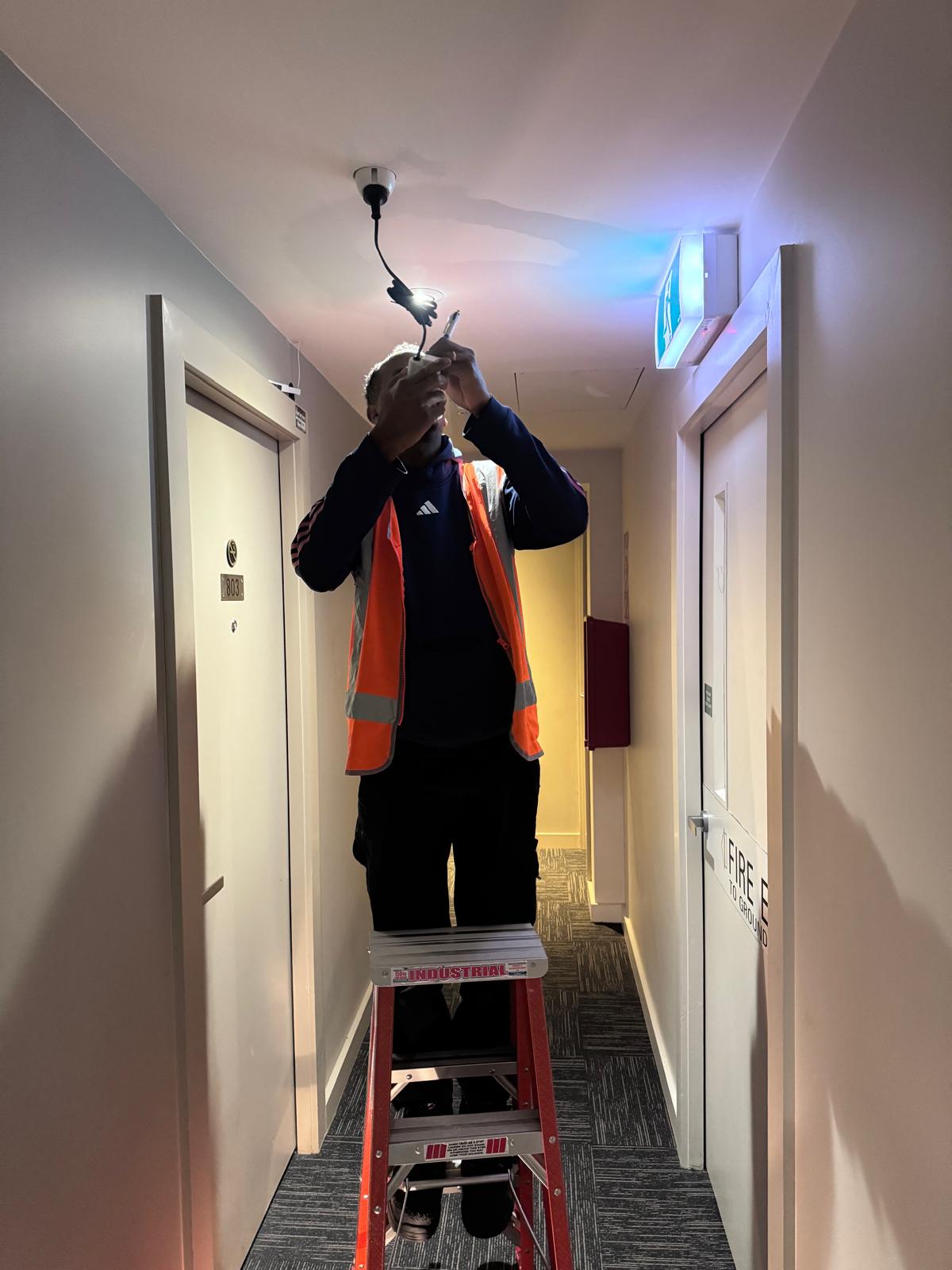 H&M Electrics electrician on a ladder installing an emergency exit light in a building corridor